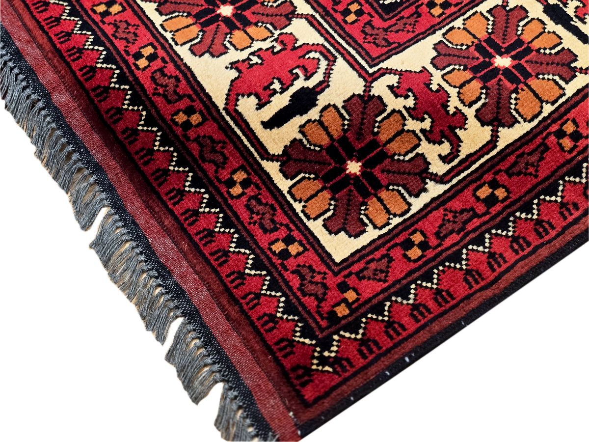 Corner view of Afghan rug with cream and red motifs; navy border and gray fringes.